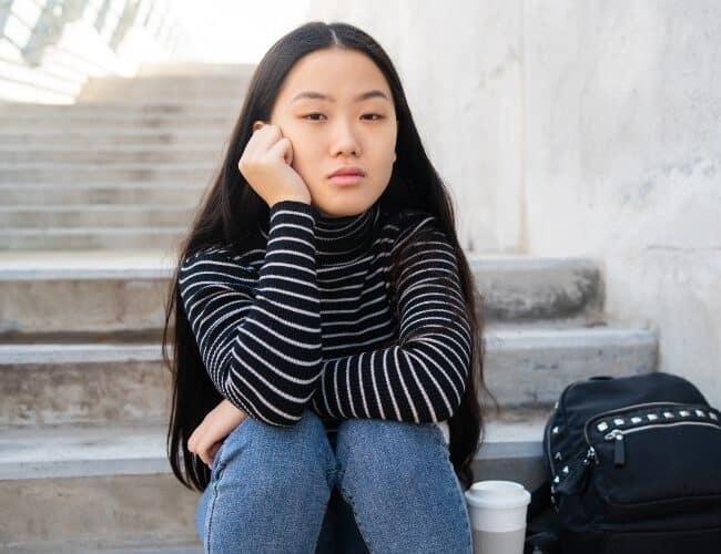 Portrait of young Asian woman with thoughtful expression sitting on concrete stairs outdoors.