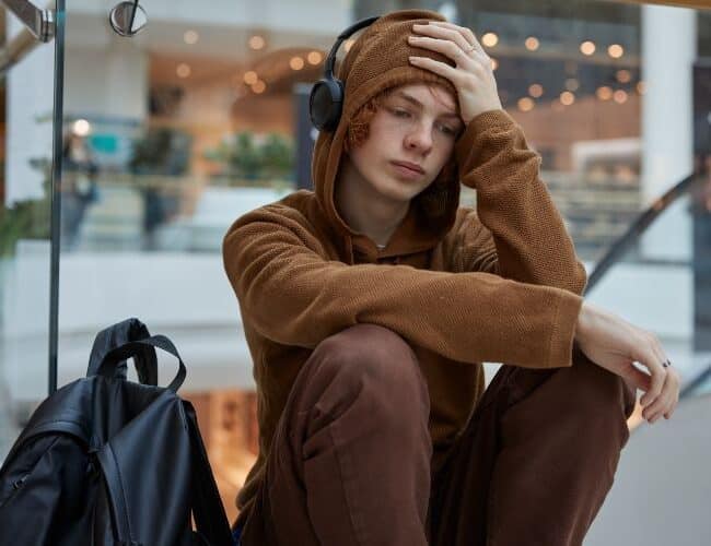 Upset teenage boy in headphones sitting nearby moving staircase at shopping mall. Depressed serious guy listening to music feeling negative emotion